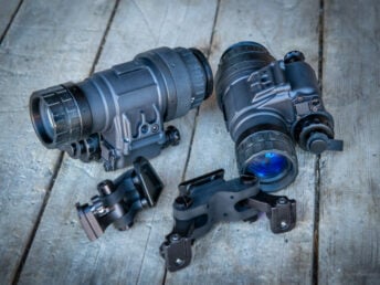 Two black night vision devices with mounting brackets on a wooden surface