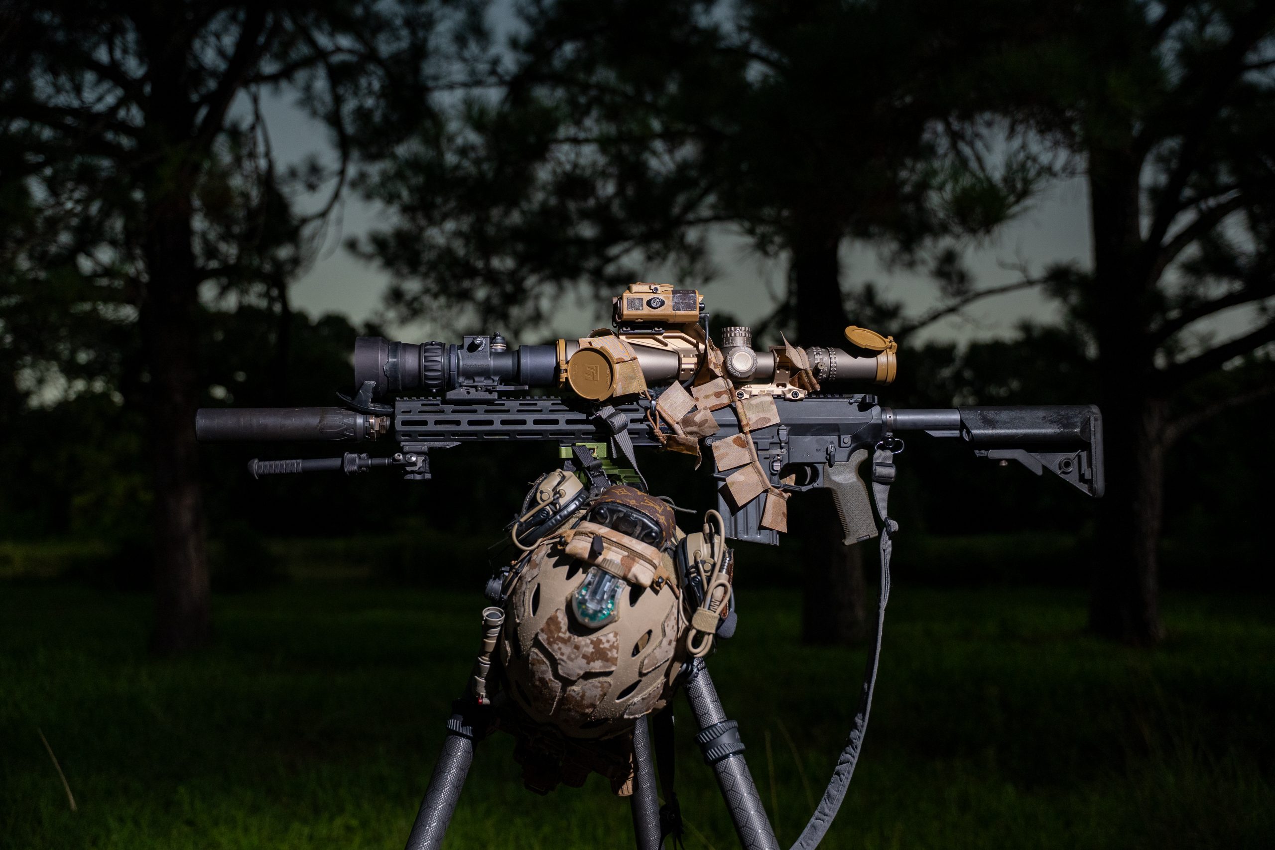 Rifle with night vision scope and tactical helmet on tripod outdoors at dusk.